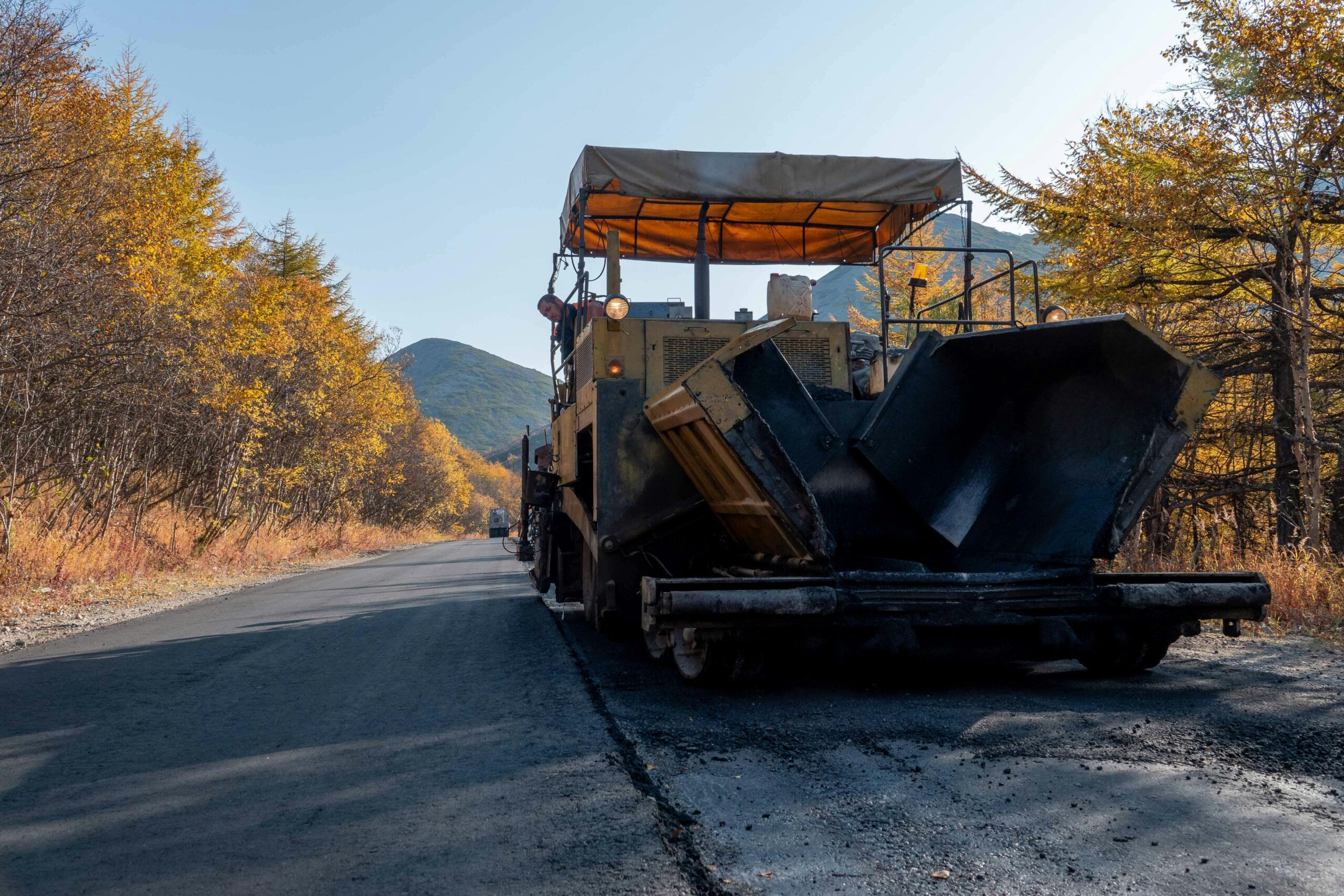 Road paving machine on a scenic autumn highway surrounded by colorful trees.